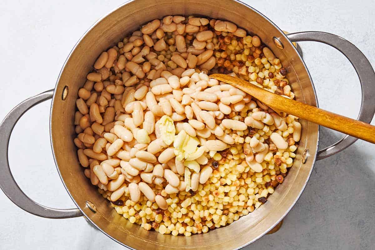 An overhead photo of fregola sarda pasta in a pot with cannellini beans, olive oil and lemon juice in a large pot with a wooden spoon.