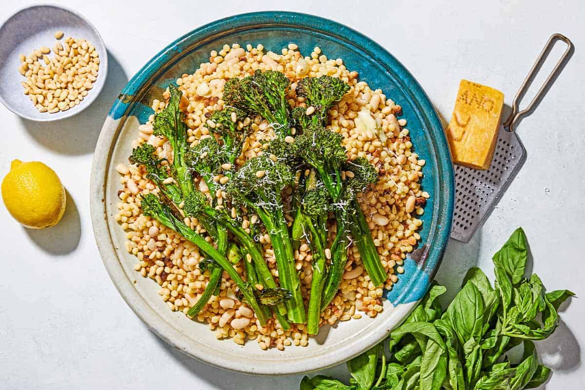 An overhead photo of fregola sarda with broccolini on a platter with a spoon. Next to this is a block of parmesan cheese, a cheese grater, some basil, a lemon and a small bowl of pine nuts.