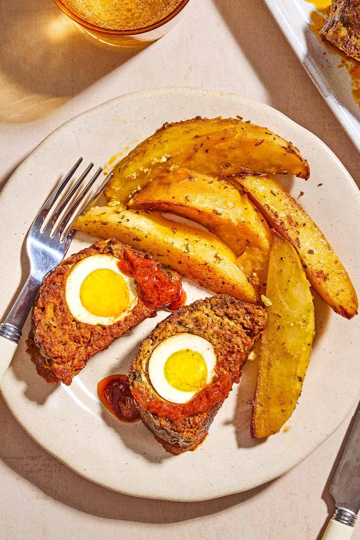 Two slices of the greek meatloaf with hard boiled eggs and sauce on a plate with greek potato wedges and a fork.