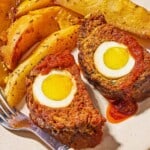 A close up of two slices of the greek meatloaf with hard boiled eggs and sauce on a plate with greek potato wedges and a fork.