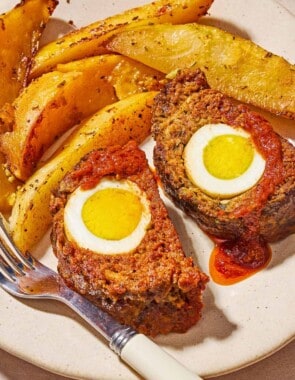 A close up of two slices of the greek meatloaf with hard boiled eggs and sauce on a plate with greek potato wedges and a fork.