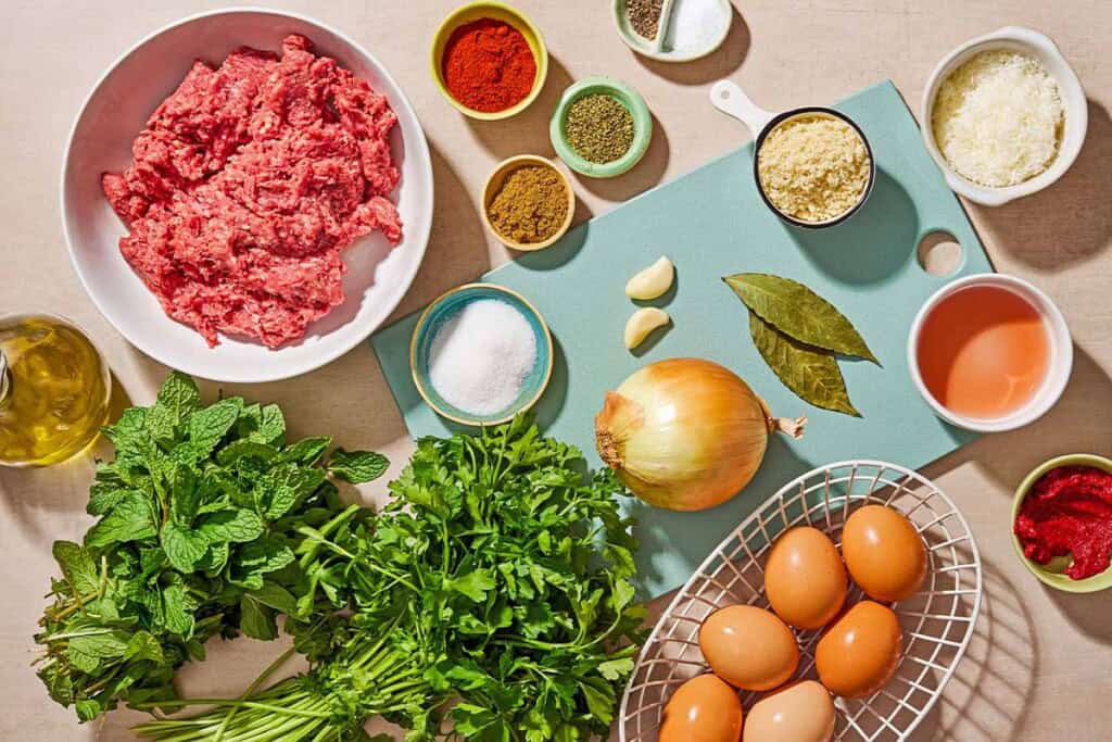 Ingredients for a greek meatloaf recipe including ground beef, eggs, onion, garlic, parsley, mint, breadcrumbs, parmesan cheese, cumin, paprika, salt, pepper, oregano, red wine vinegar, olive oil, tomato paste bay leaves and sugar.