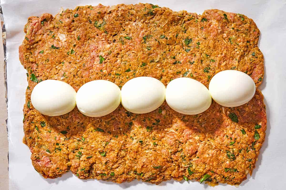 The meatloaf mixture for the greek meatloaf flattened into the shape of a rectangle with a row of 5 hard boiled eggs going down the middle on a parchment lined sheet of aluminum foil.