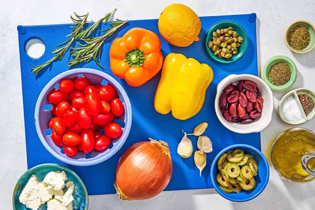 Ingredients for Mediterranean vegetable medley including bell peppers, lemon, onion, garlic, grape tomatoes, feta, olive oil, fresh rosemary, greek oregano, dried thyme, ground black pepper, salt, kalamata olives, castelventrano olives, and capers.