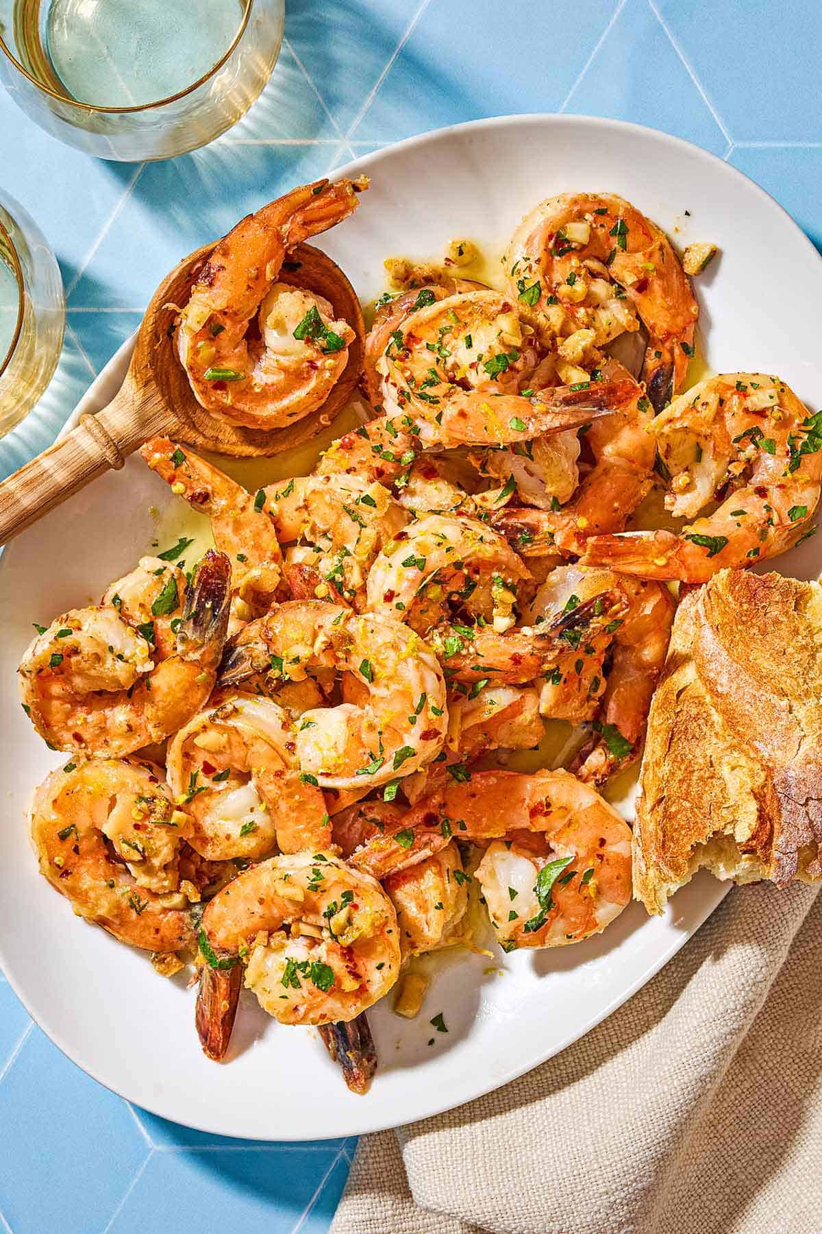 An overhead photo of shrimp scampi on a platter with a wooden spoon and a piece of crusty bread. Next to this is a cloth napkin and 2 glasses of wine.