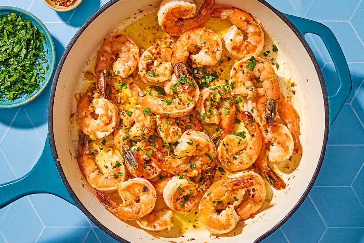 An overhead photo of shrimp scampi in a skillet next to a bowl of chopped parsley.