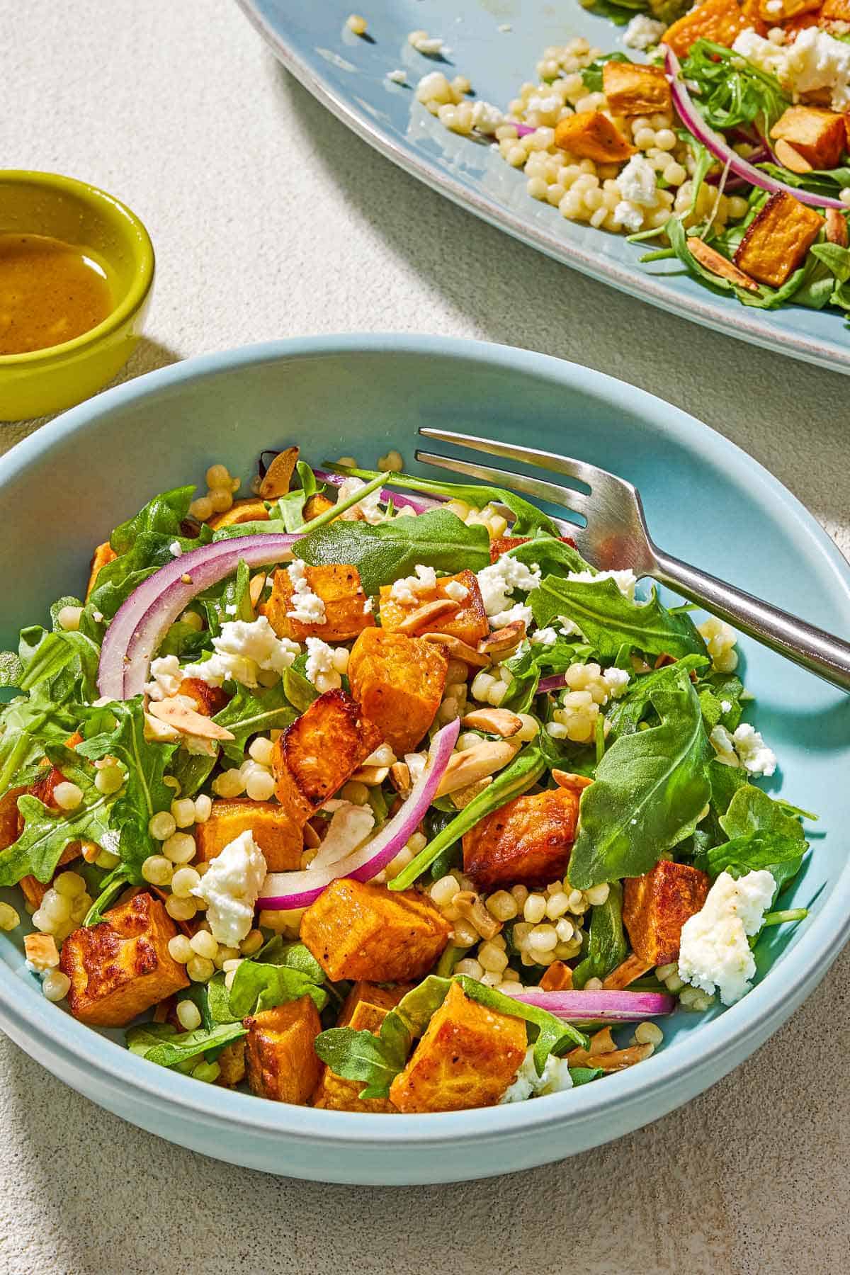 A serving of the sweet potato salad in a bowl with a fork next to a small bowl of the the dressing and the rest of the salad on a platter.