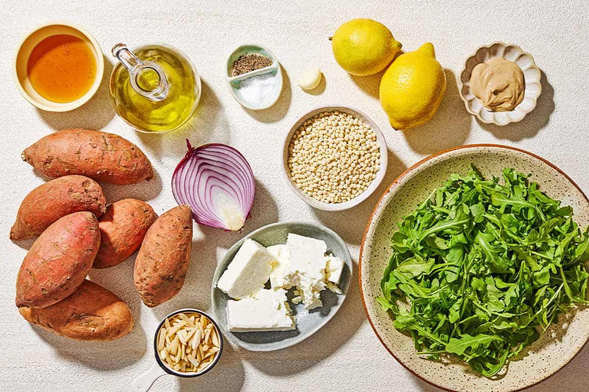 Ingredients for sweet potato salad including sweet potatoes, olive oil, salt, black pepper, arugula, red onion, feta cheese, slivered almonds, honey, lemons, garlic, and pearl couscous.