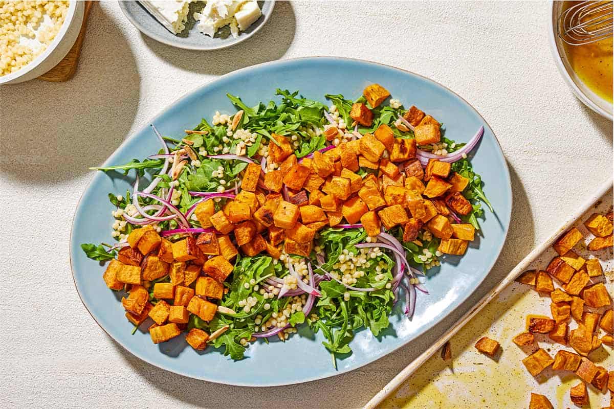 The sweet potato salad partially assembled on a platter next to the roasted sweet potato cubes on a sheet pan, the dressing in a bowl with a whisk, some crumbled feta cheese and a bowl of cooked pearl couscous.