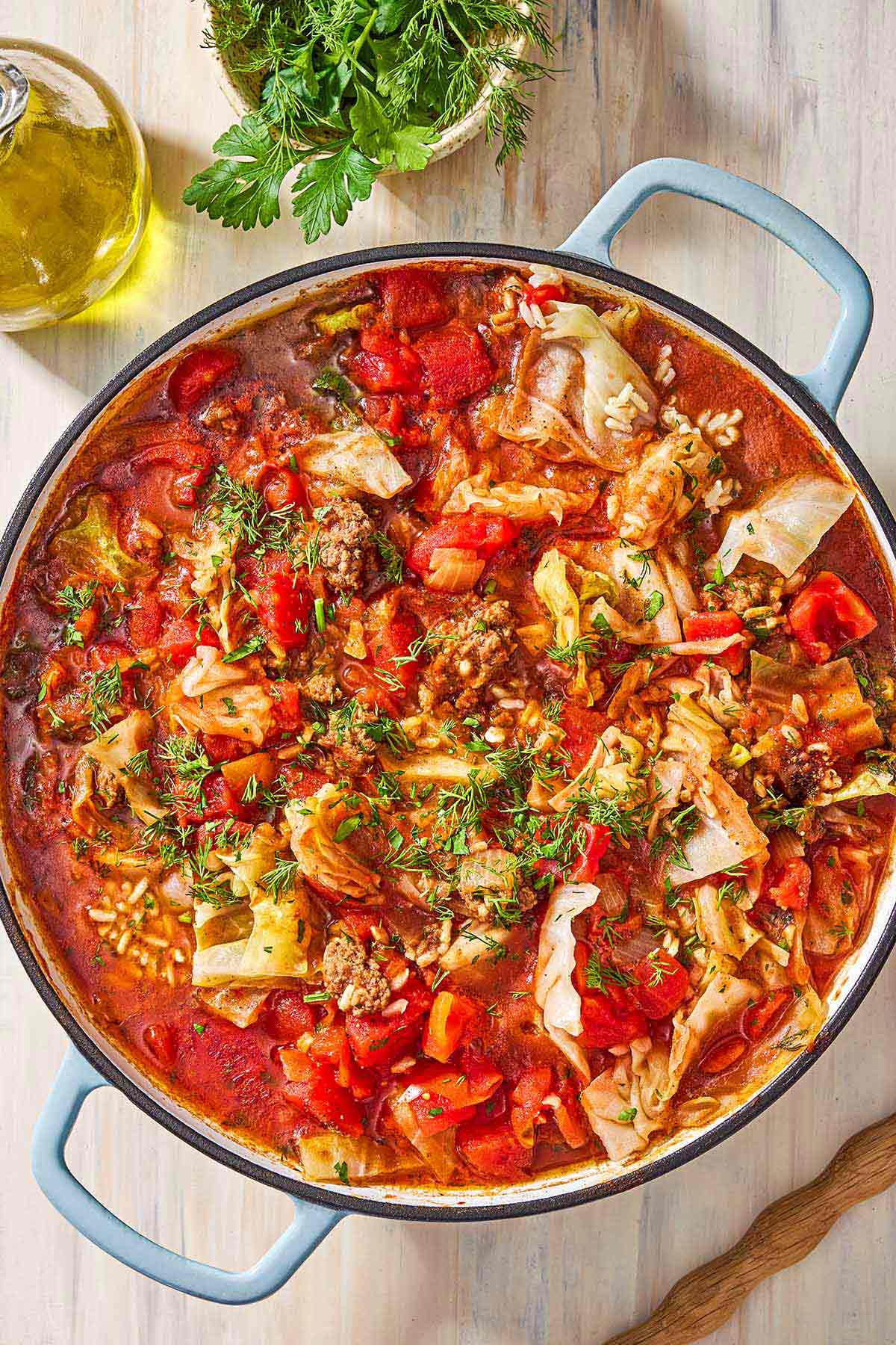 An overhead photo of unstuffed cabbage rolls in a skillet. Next to this is a small bowl of Italian parsley and dill, and a jar of olive oil.