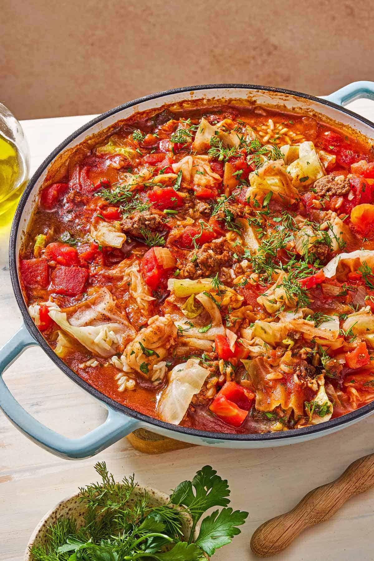 A close up of unstuffed cabbage rolls in a skillet. Next to this is a small bowl of Italian parsley and dill.