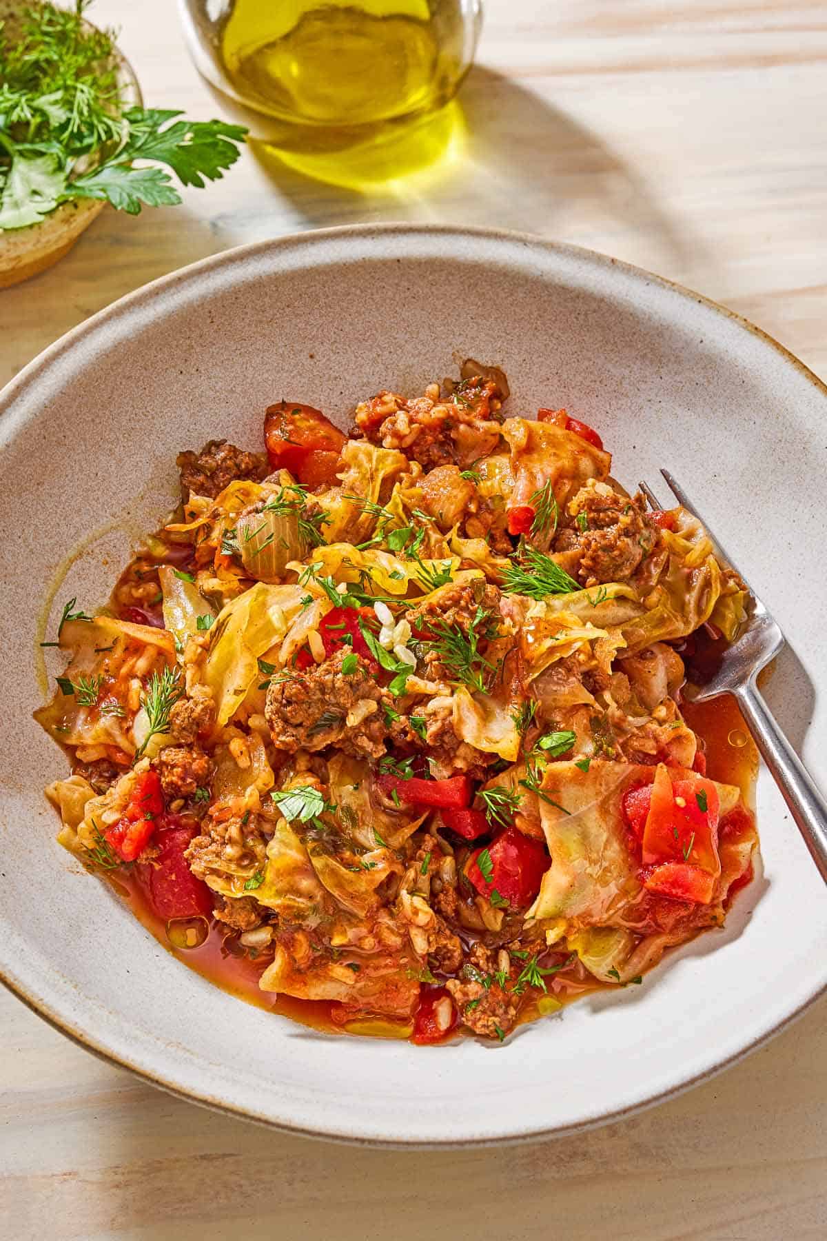 A close up of a serving of the unstuffed cabbage rolls in a bowl with a fork. Next to this is a bowl with dill and parsley, and a jar of olive oil.