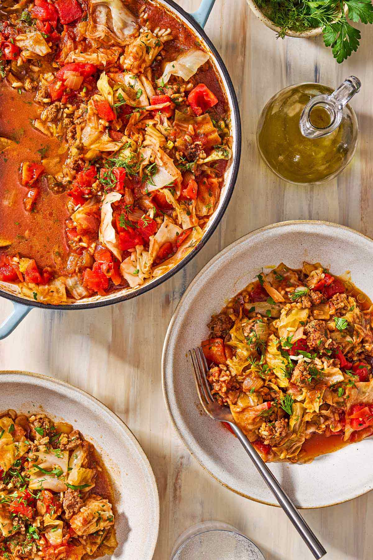 An overhead photo of a serving of the unstuffed cabbage rolls in a bowl with a fork. Next to this is another serving in a bowl, the rest of the unstuffed cabbage rolls in a skillet, a jar of olive oil, and a glass of sparkling water.