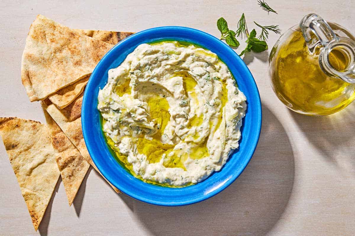 Vegan tzatziki sauce with a drizzle of olive oil in a bowl next to pita wedges and a small jug of olive oil.