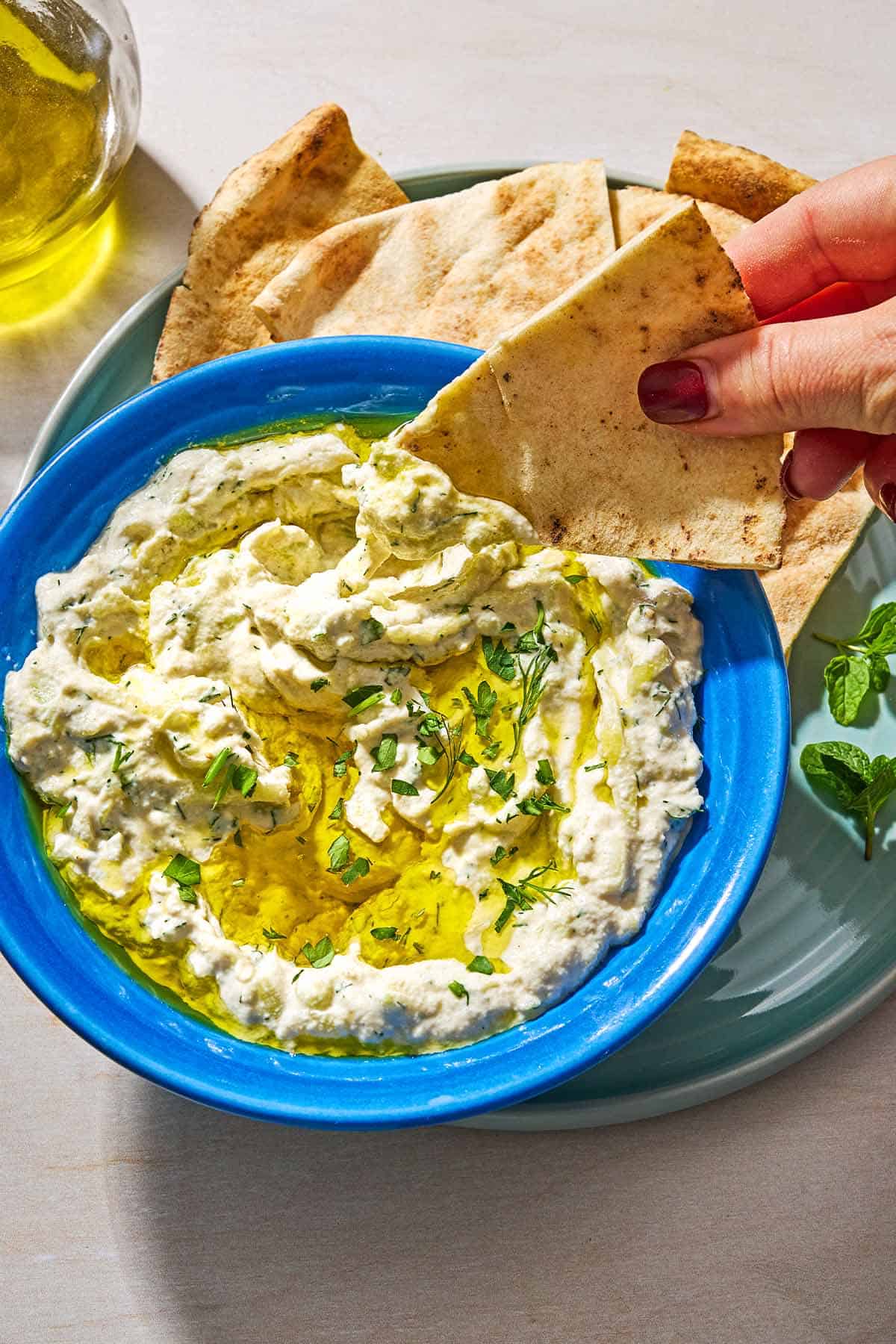 A pita wedge being dipped into a bowl of dairy free tzatziki that is on a plate with more pita wedges.