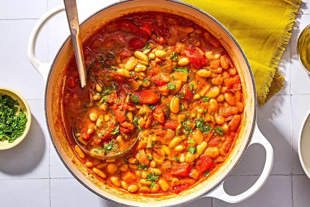 White bean stew in a large pot with a ladle next to a cloth napkin and a bowl of chopped parsley.