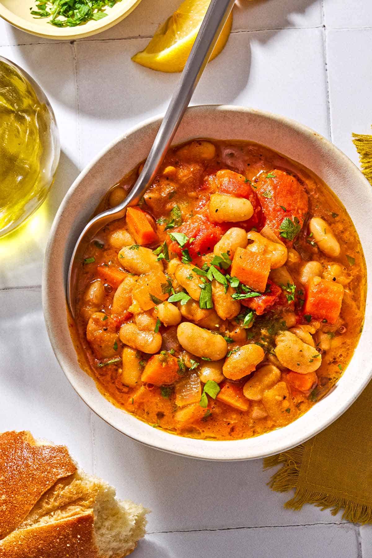 White bean stew in a bowl surrounded by a cloth napkin, some crusty bread, and a lemon wedge.