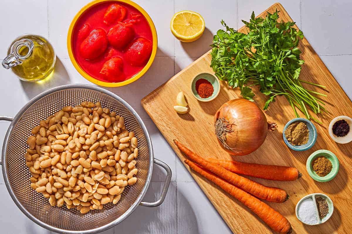 Ingredients for white bean stew including cannellini beans, canned whole tomatoes, lemon, garlic, olive oil, carrots, onion, parsley, salt, pepper, urfa biber, cumin, paprika, and coriander.