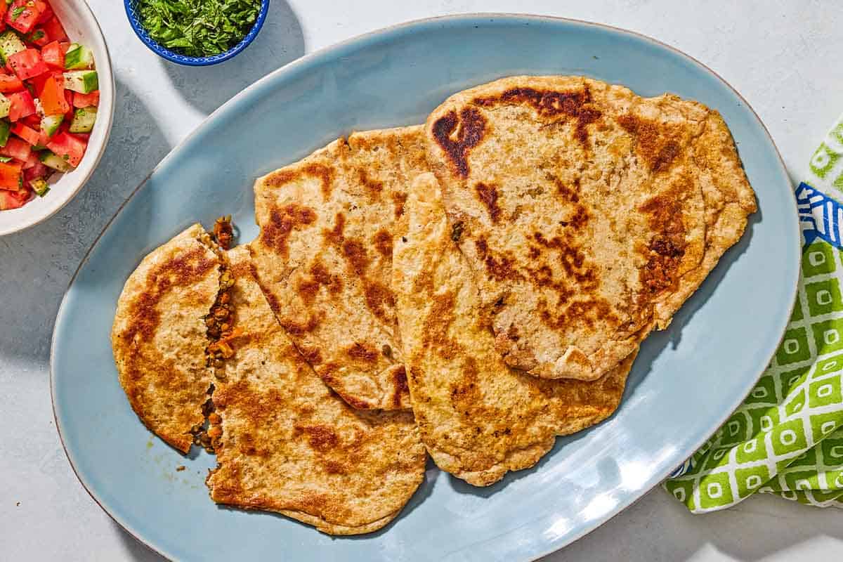 A blue platter is resting near a green and blue napkin. Whole wheat stuffed flat breads are on the platter and one is torn open showing a lentil and carrot spiced filling. 