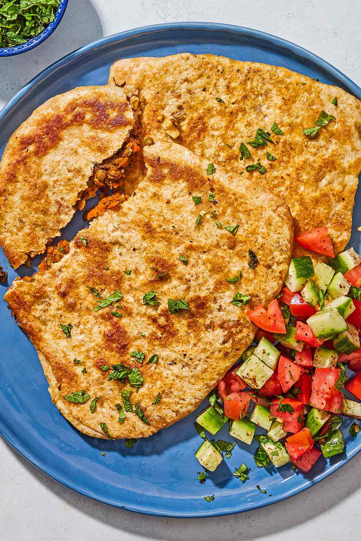 A blue plate is resting near a green and blue napkin. Whole wheat stuffed flat breads are on the platter and one is torn open showing a lentil and carrot spiced filling. Next to it is a cucumber and tomato salad. 