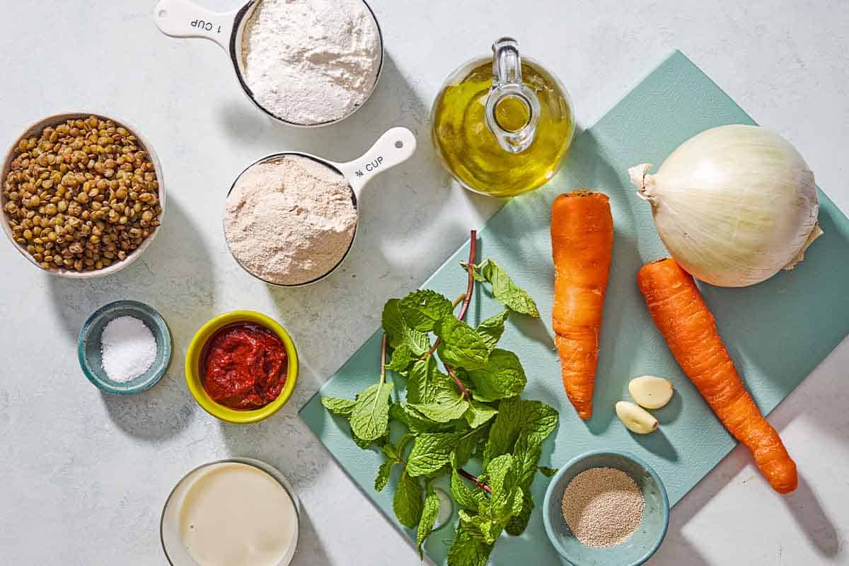 Carrots, lentils, mint, harrissa, flour, non-dairy milk and onion on a blue cutting board and white counter. 