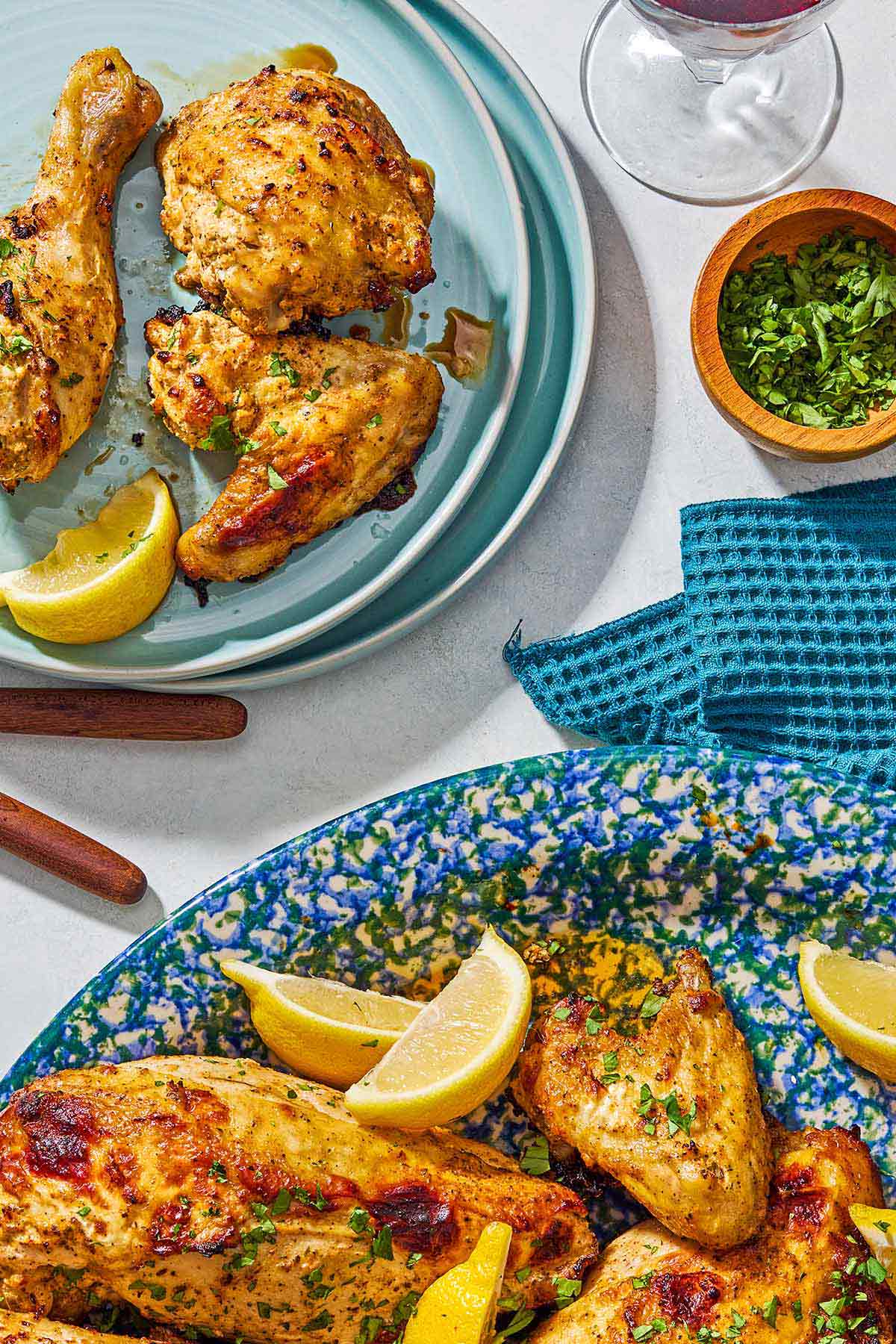 3 pieces of cooked Greek yogurt marinated chicken on a plate with a lemon wedge next to a glass of wine, a bowl of chopped parsley, a kitchen towel and the rest of the yogurt chicken with lemon wedges on a platter.