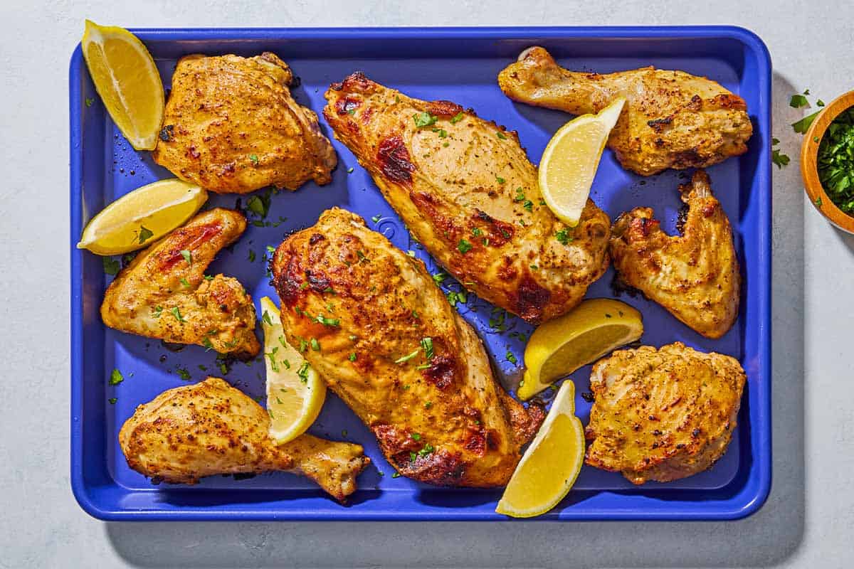 Pieces of cooked Greek yogurt marinated chicken on a sheet pan with lemon wedges next to a bowl of chopped parsley.