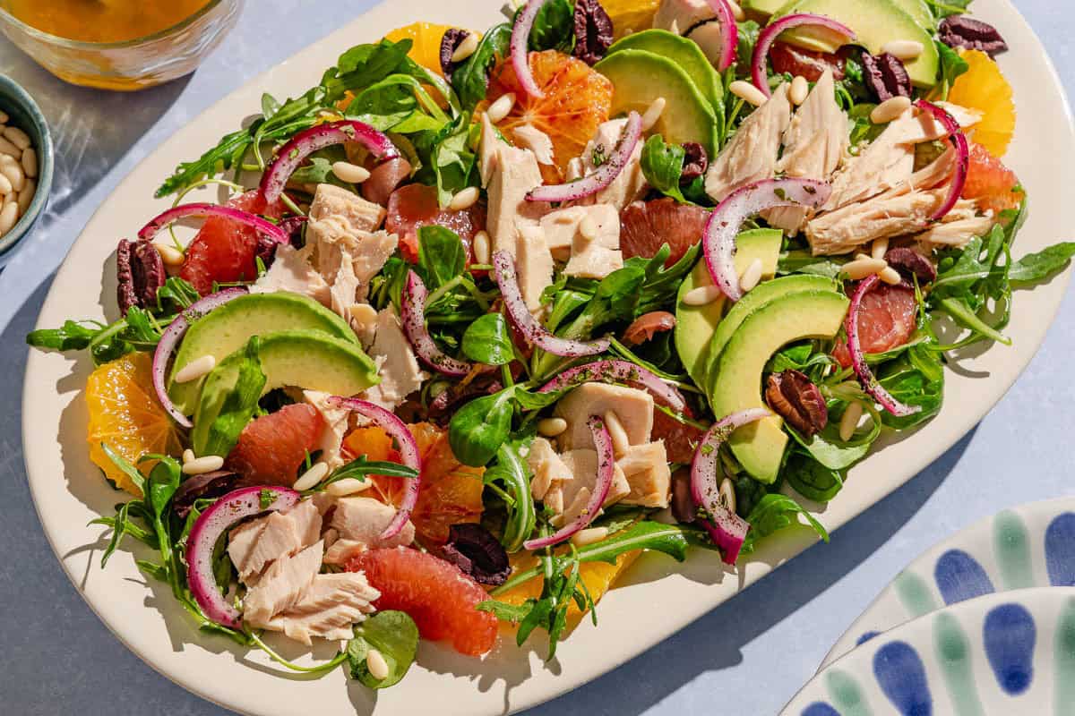 Avocado tuna salad on a platter surrounded by a stack of 2 plates, salad dressing in a bowl, and a bowl of pine nuts.