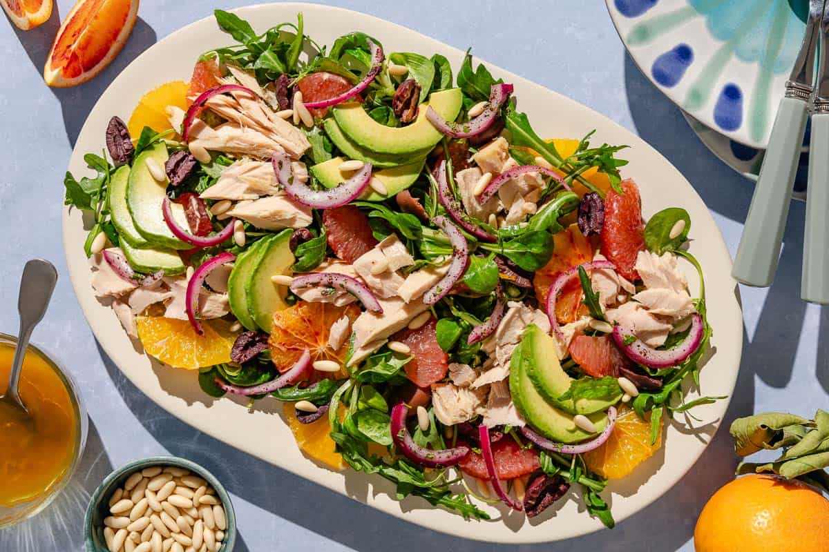 Avocado tuna salad on a platter surrounded by a stack of 2 plates with forks, orange wedges, salad dressing, and a bowl of pine nuts.