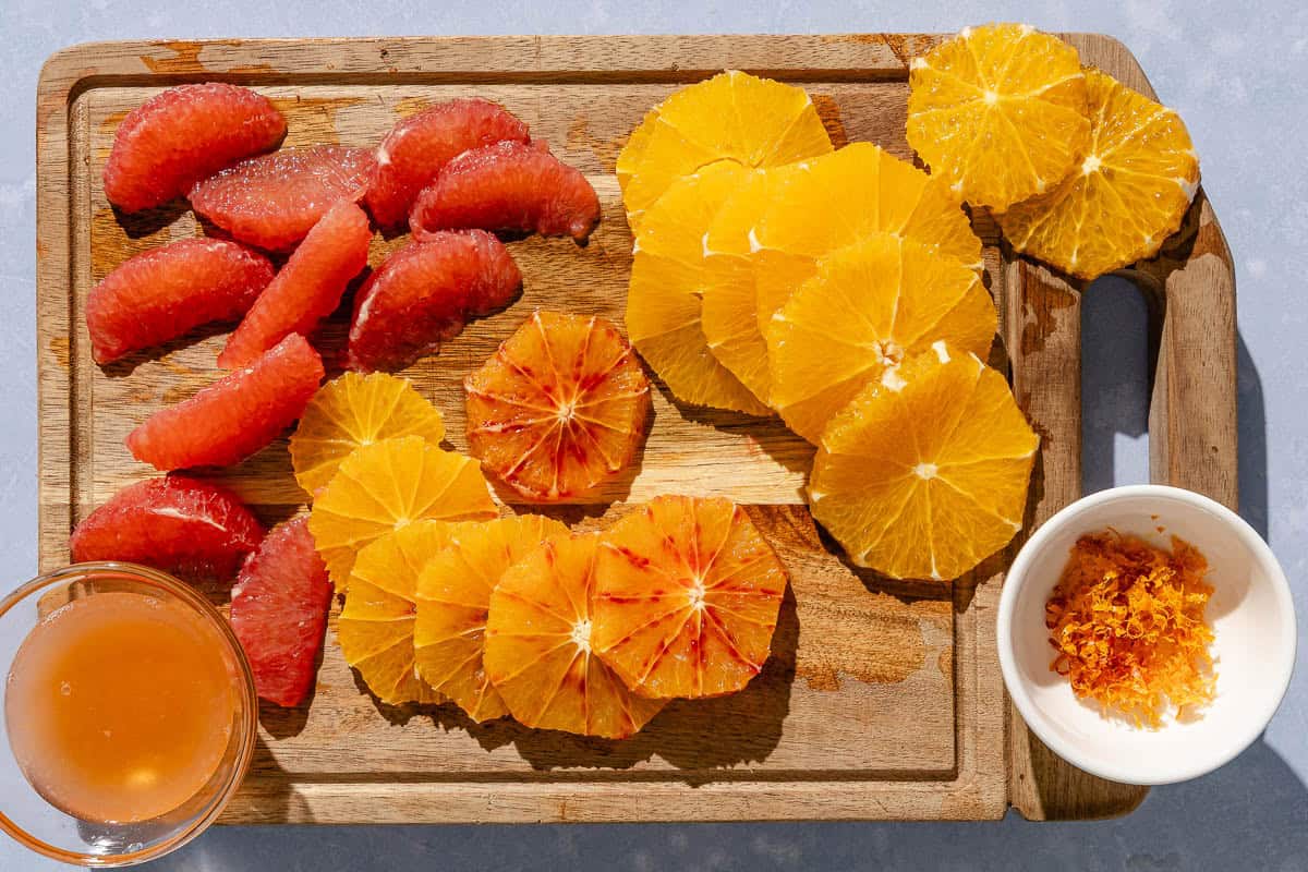 Peeled and sliced oranges and grapefruit on a cutting board with a bowl of zest and a bowl of juice.