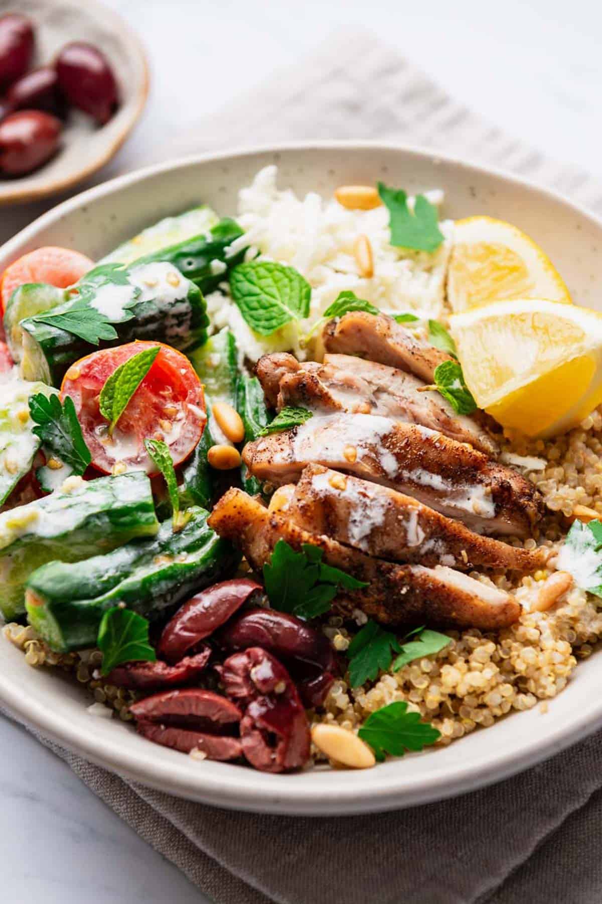 A quinoa chicken bowl on resting on a cloth napkin with a small bowl of kalamata olives nearby.