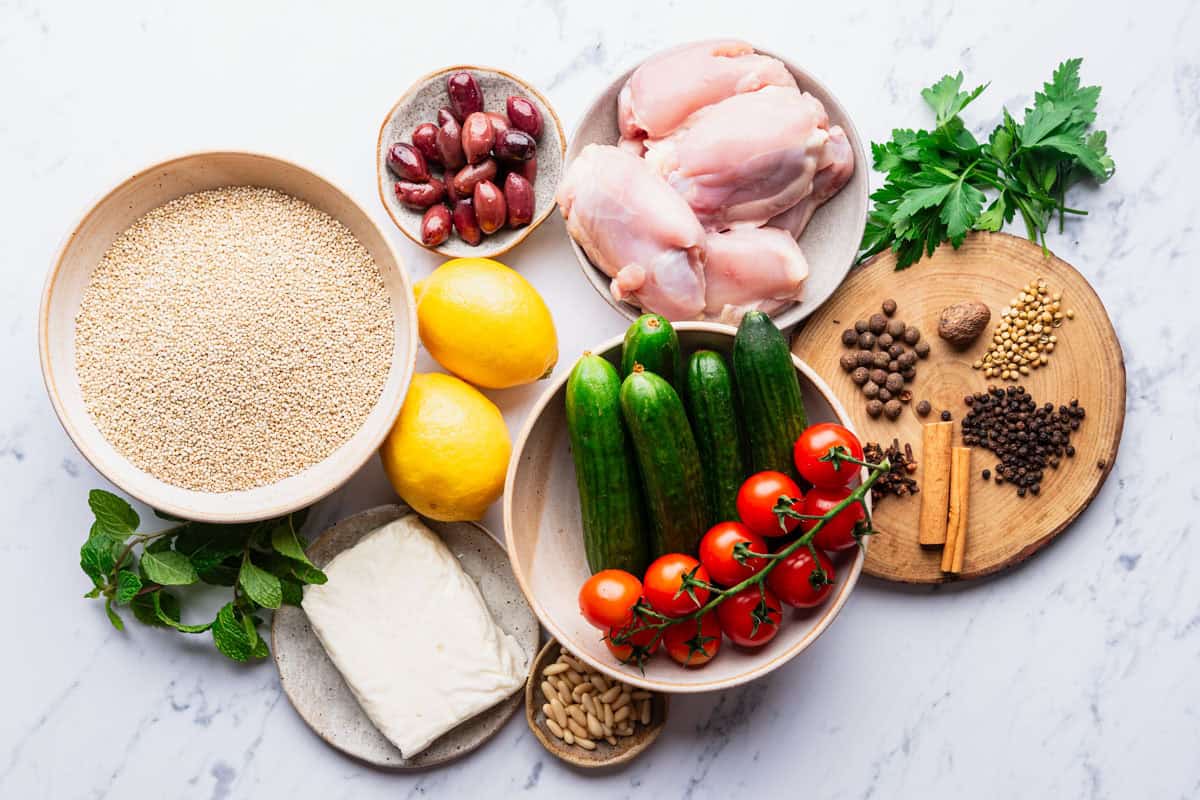 Ingredients for the chicken quinoa bowl recipe including allspice, cinnamon, black pepper, coriander, nutmeg, cloves, persian cucumbers, cherry tomatoes, salt, quinoa, greek yogurt, lemon, boneless skinless chicken thighs, olive oil, halloumi, pine nuts, mint, parsley, kalamata olives and lemons.