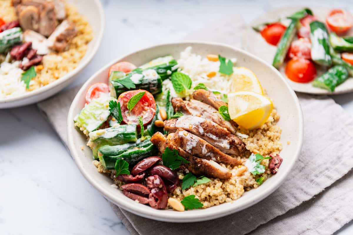 A quinoa chicken bowl on resting on a cloth napkin with a plate of the cherry tomato and smashed cucumber topping and another quinoa chicken bowl nearby.