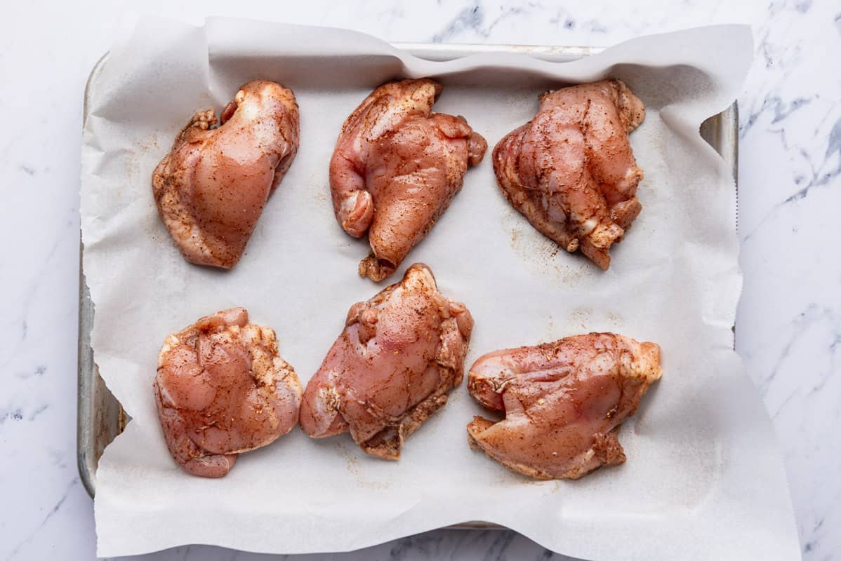 Seasoned boneless skinless chicken thighs spread evenly on a parchment lined sheet pan.