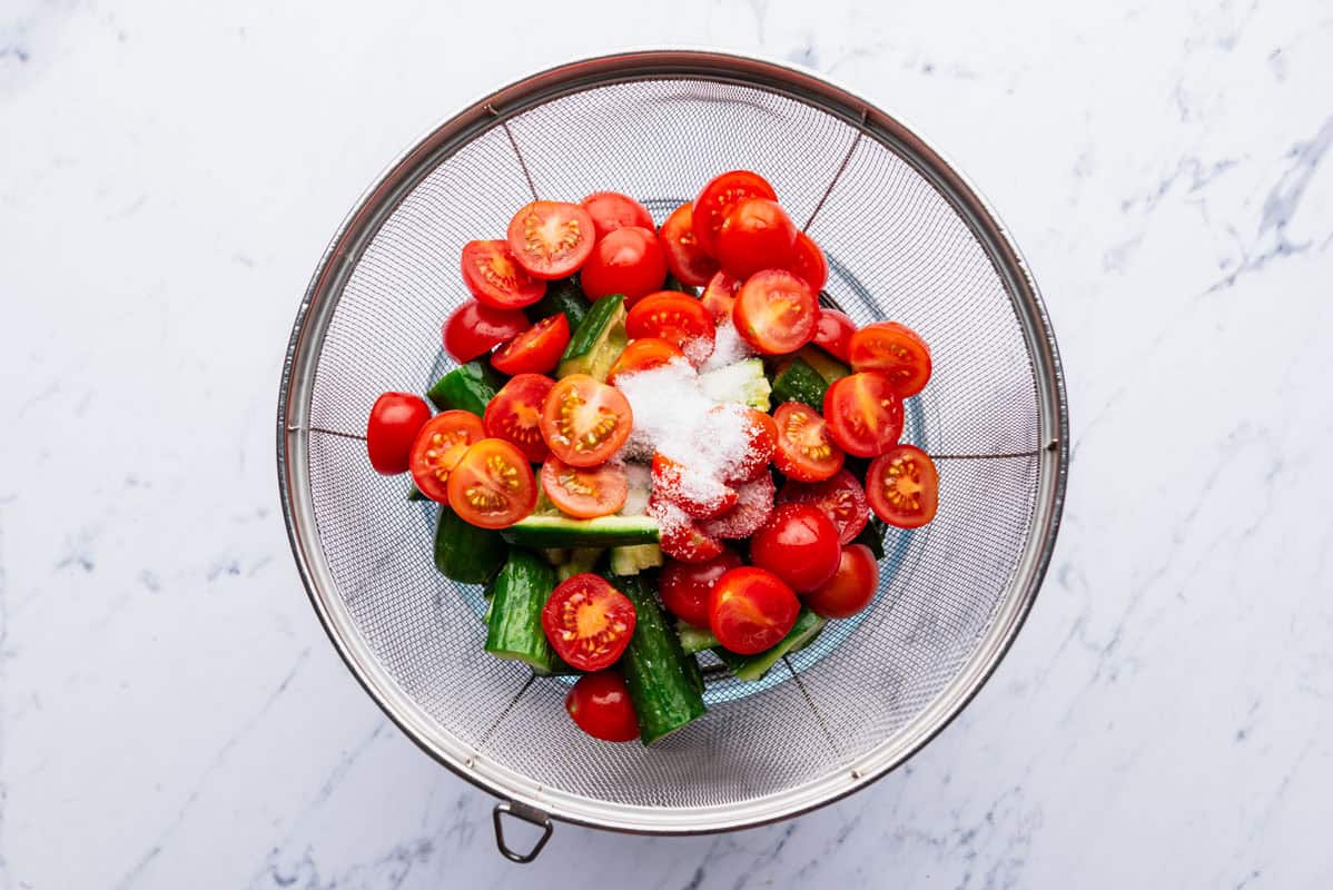 Cherry tomato halves and smashed cucumbers topped with salt in a strainer.