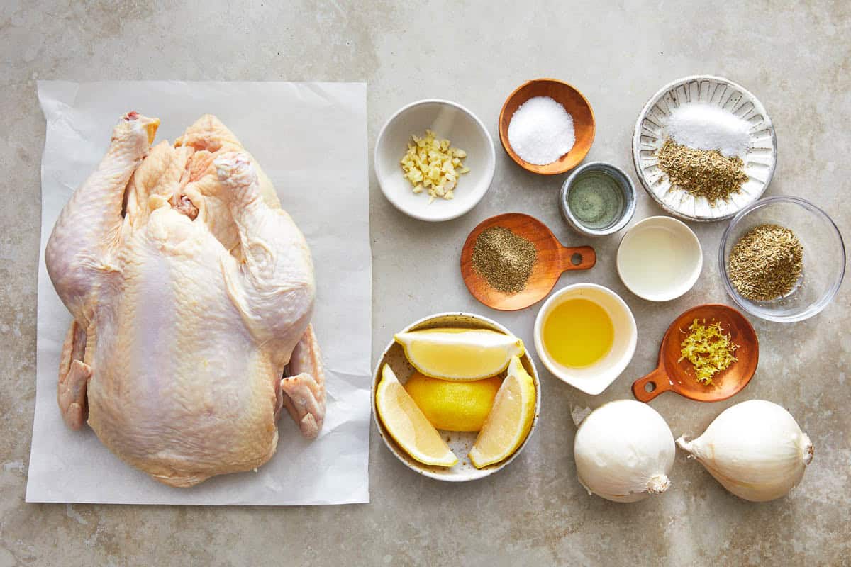 Ingredients for roasted whole chicken including a chicken, garlic, lemon, onions, olive oil and spices.