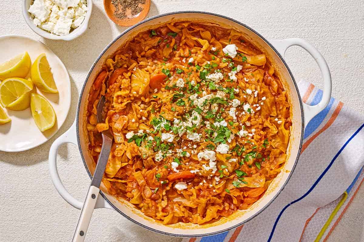 Greek cabbage rice topped with crumbled feta and chopped parsley in a large pot with a serving spoon next to a kitchen towel, a plate of lemon wedges, and bowls of crumbled feta and black pepper.