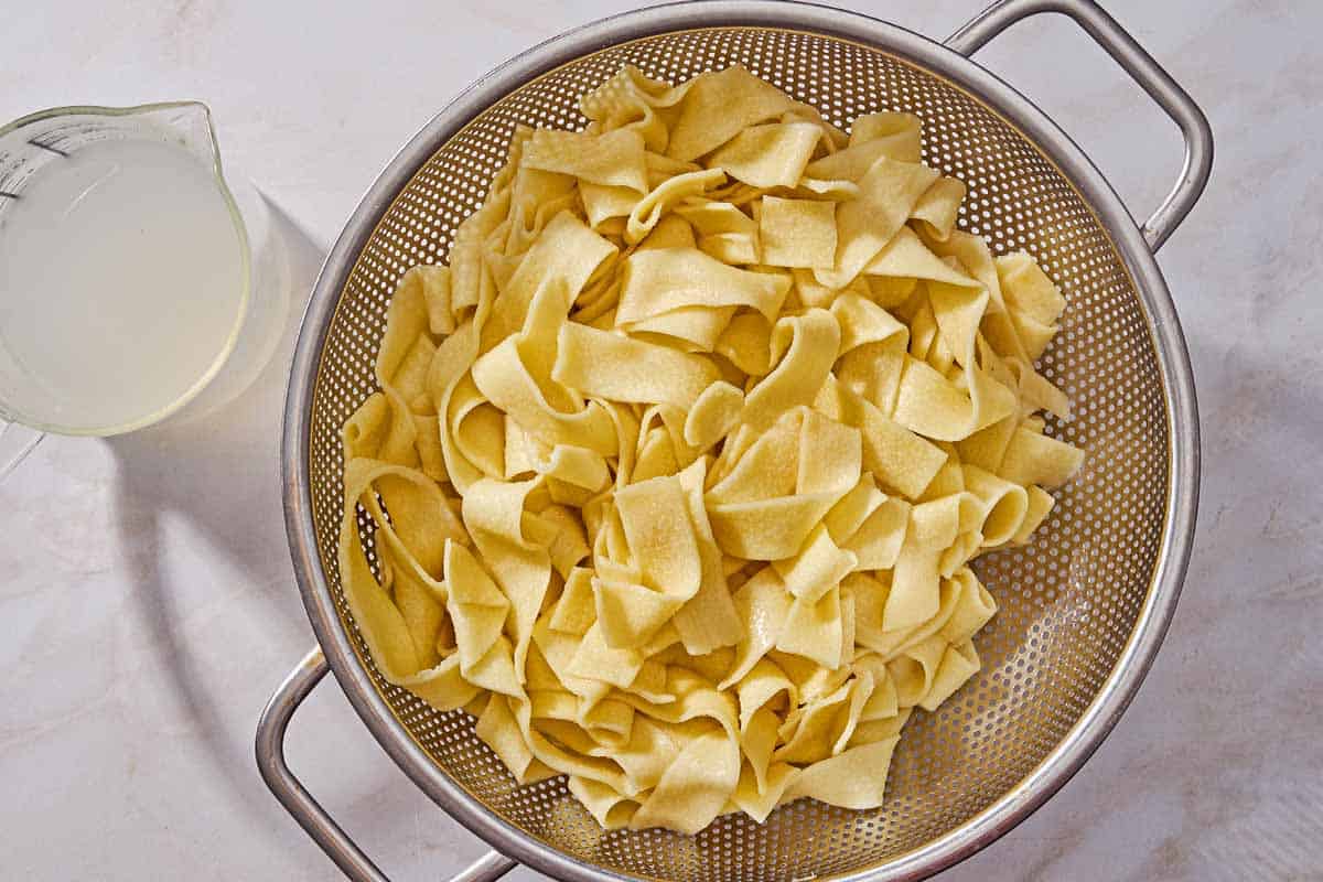 Cooked pasta in a strainer next to a measuring cup with pasta water.