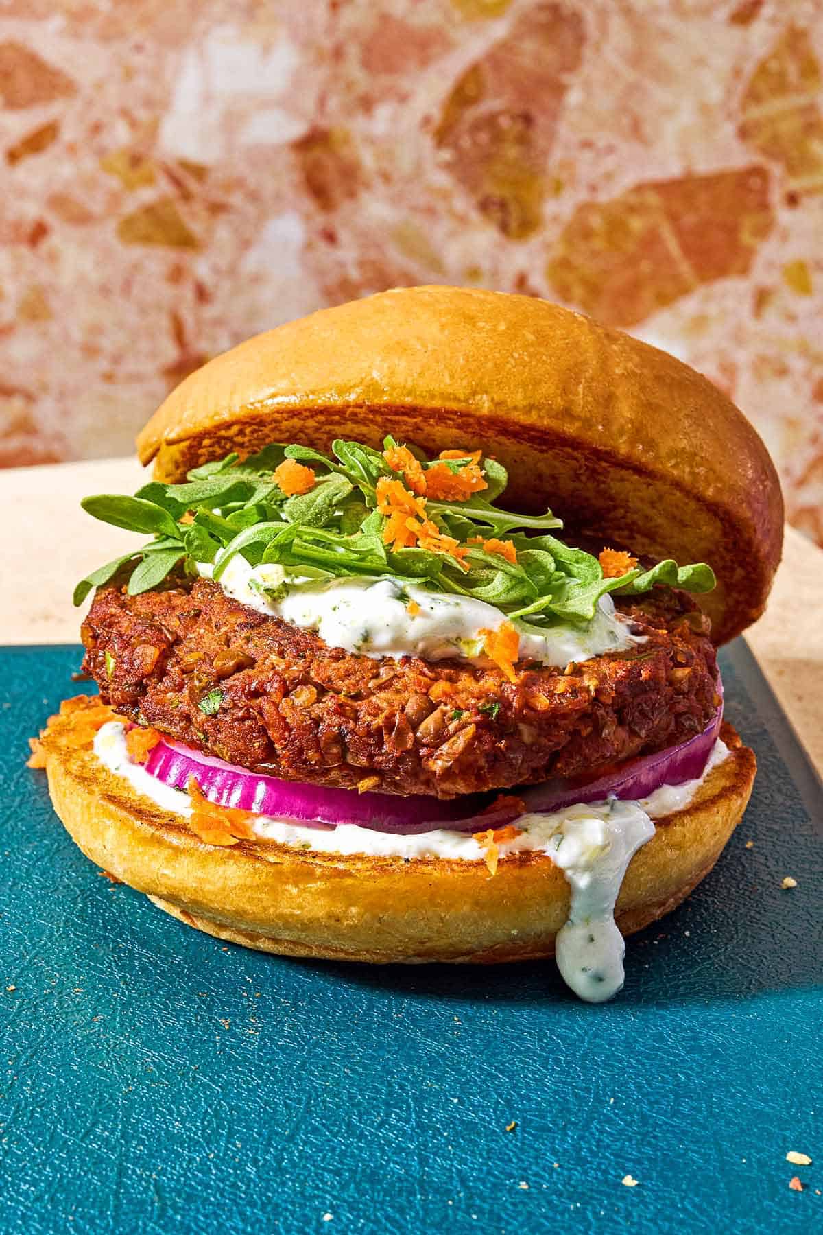 A vegetarian burger on a bun with yogurt herb sauce, red onion, grated carrots and arugula leaves.