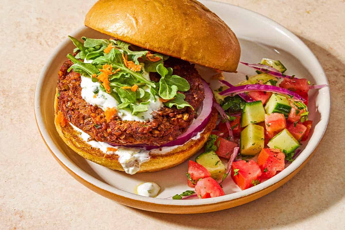 A plate with a cucumber tomato salad and a vegetarian burger on a bun with yogurt herb sauce, red onion, grated carrots and arugula leaves.