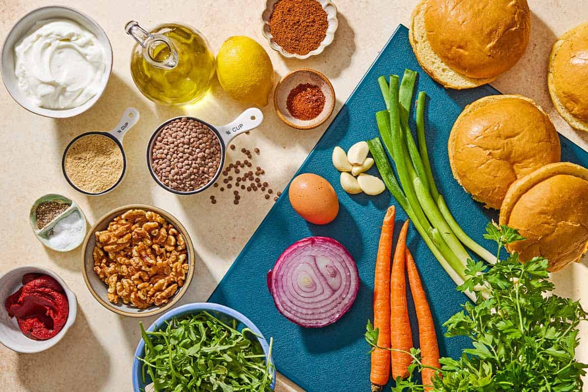 Ingredients for lentil burgers including brown lentils, walnuts, breadcrumbs, green onions garlic, tomato paste, aleppo pepper, baharat, salt, black pepper, egg, olive oil, Greek yogurt, parsley, lemon, garlic, buns, carrots, red onion and arugula leaves.