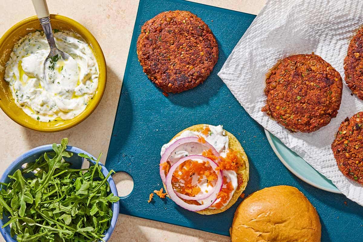 4 lentil burgers on a cutting board with a bun topped with grated carrots, red onion and yogurt herb sauce next to bowls of arugula and the rest of sauce in a bowl with a spoon.