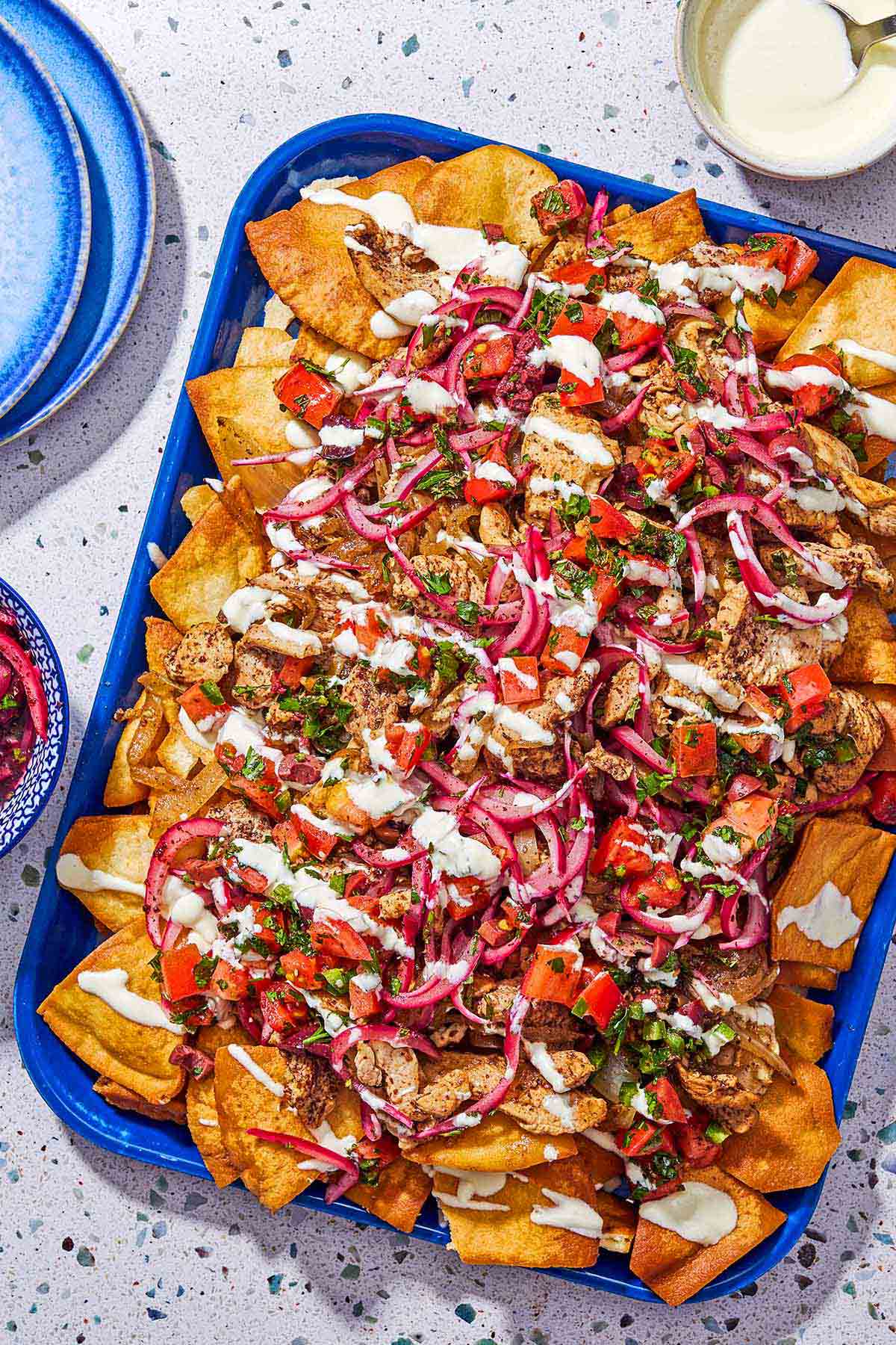 Healthy nachos on a platter surrounded by a stack of 2 plates and a bowl of feta dressing with a spoon.