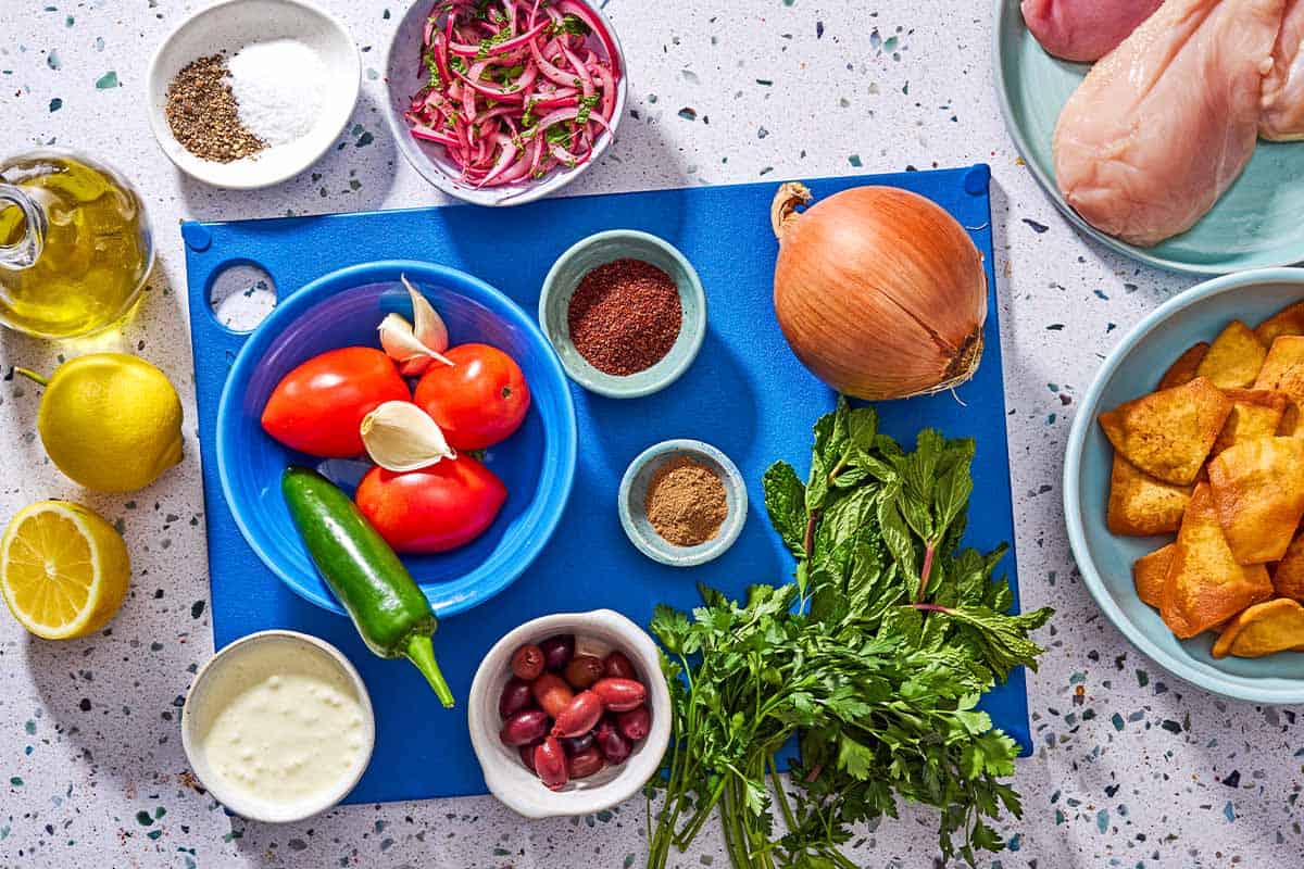 Ingredients for chicken nachos including tomatoes, jalapeno, kalamata olives, parsley, mint, sumac, salt, black pepper, lemon, olive oil, onion, garlic, chicken, and allspice.