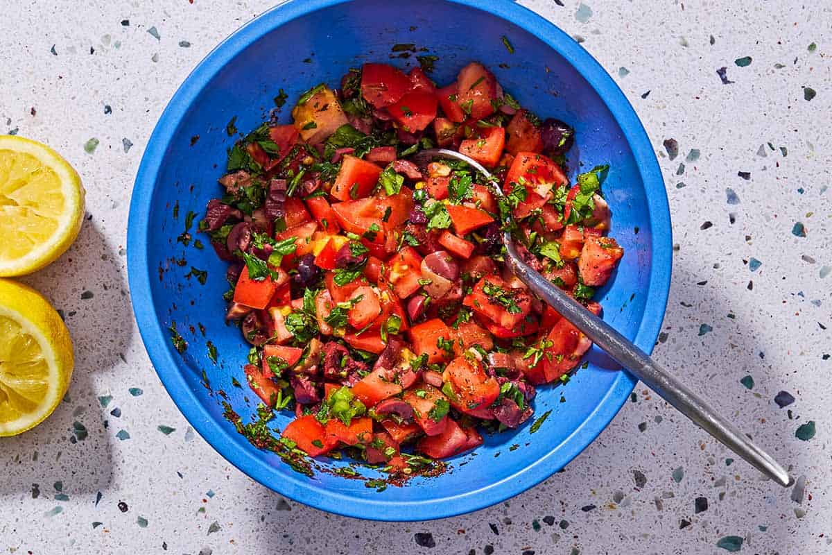 Mediterranean salsa in a bowl with a spoon next to 2 juiced lemon halves.