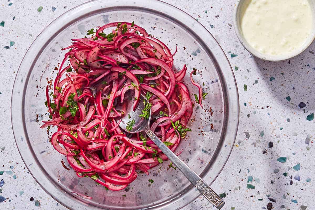 Pickled red onions in a bowl with a spoon next to a bowl of feta dressing.