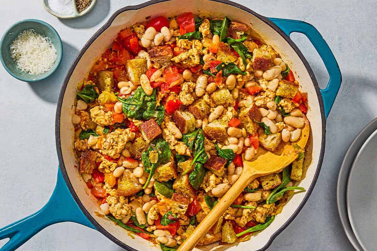 The ground turkey recipe in a skillet with a wooden spoon next to a stack of two plates a bowl with salt and black pepper, and a small bowl of grated parmesan cheese.