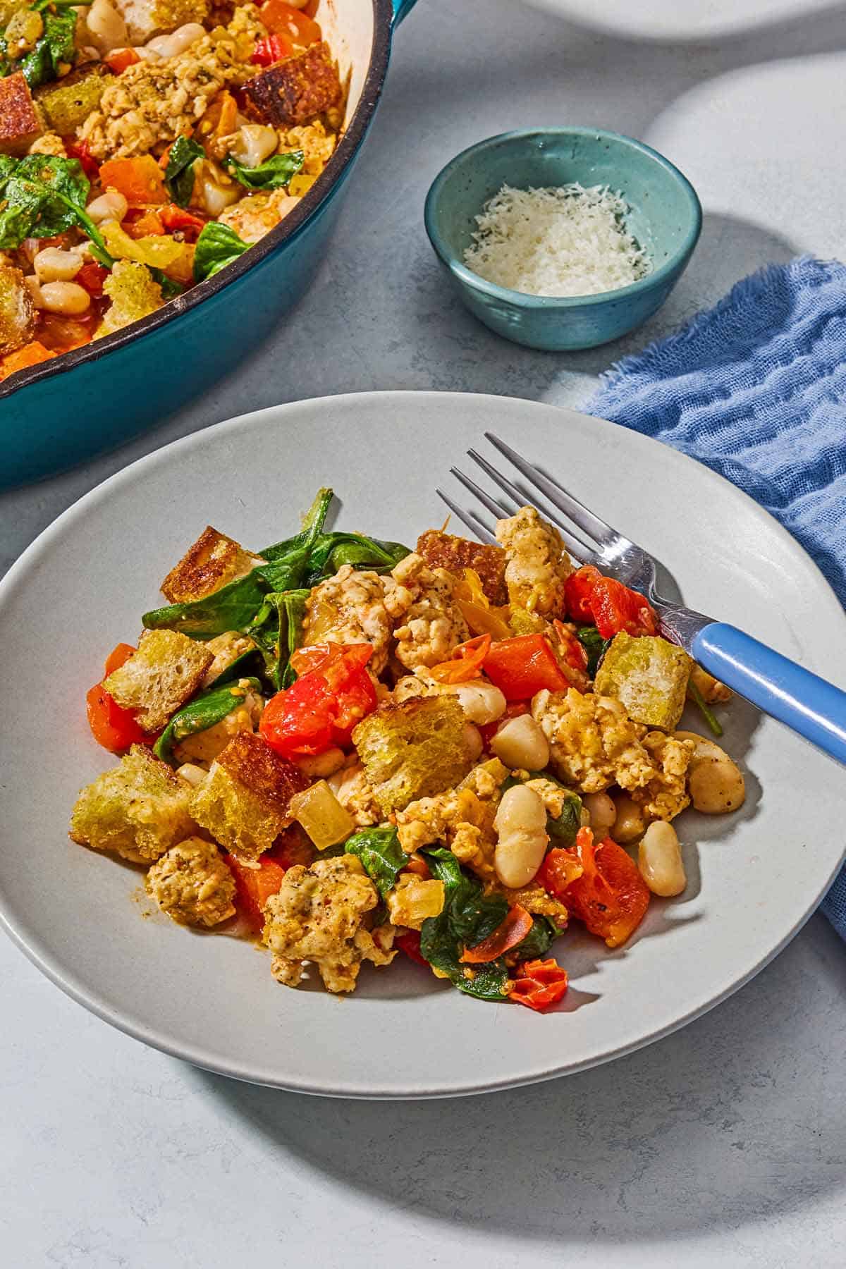 A serving of the ground turkey recipe on a plate with a fork with a small bowl of parmesan cheese and the rest of the ground turkey recipe in a skillet in the background.