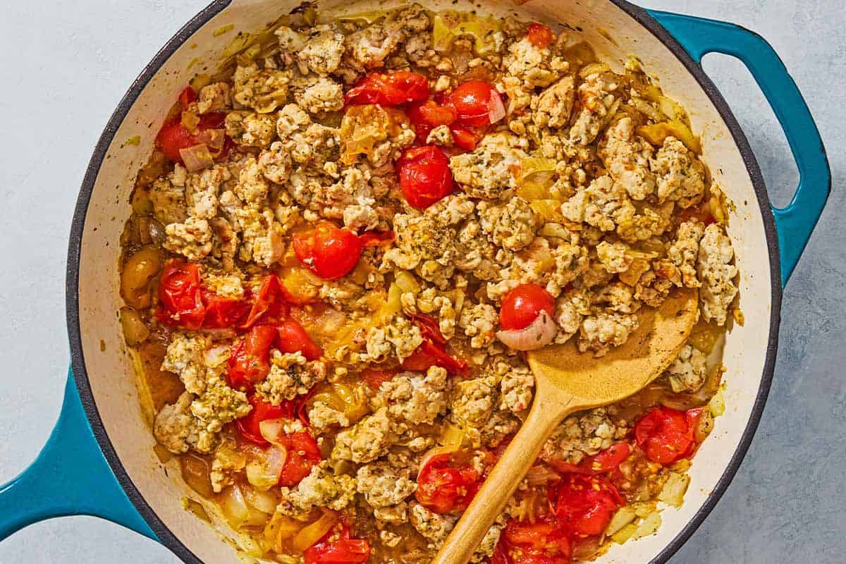 Ground turkey, tomatoes, onions and seasonings being cooked in a skillet with a wooden spoon.