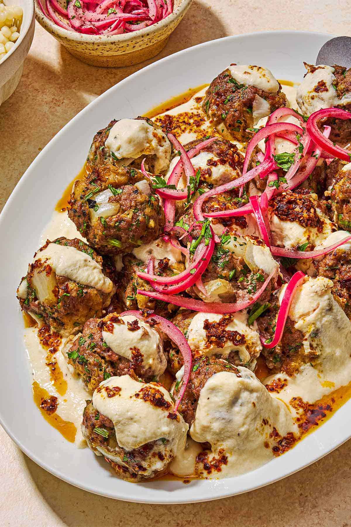 Turkish meatballs and yogurt sauce topped with pickled red onions and chopped parsley on a white platter next to a bowl pickled red onions.